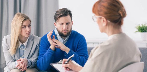 תמונת נושא של המאמר על ייעוץ זוגי לפני נישואין המציג זוג צעיר יושב בלחץ בייעוץ זוגי מול פסיכולוגית או יועצת עם שיער ג'ינג'י המקשיבה ורושמת