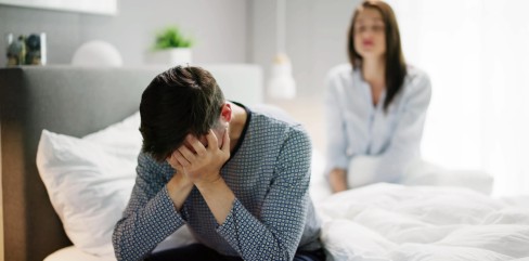 תמונת נושא של המאמר מטפלת זוגית מה חשוב לדעת לפני הפגישה הראשונה? בה רואים גבר יושב על קצה המיטה עם ראש בידיים ואישה יושבת מטושטשת ברקע