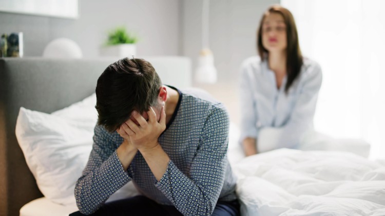 תמונת נושא של המאמר מטפלת זוגית מה חשוב לדעת לפני הפגישה הראשונה? בה רואים גבר יושב על קצה המיטה עם ראש בידיים ואישה יושבת מטושטשת ברקע