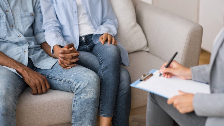 תמונת נושא של המאמר על יועצת זוגית מומלצת איך לבחור המציגה זוג צעיר יושב יחד על ספה כשהם אוחזים ידיים בג'ינס וחולצות כחולות מול מטפלת או יועצת עם לוח ונייר המייצגים טיפול זוגי