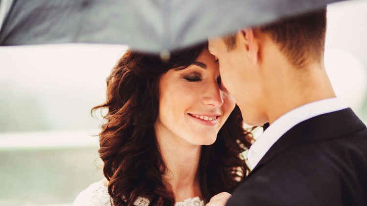 תמונת נושא של המאמר ייעוץ נישואין צילום תקריב של זוג ביום חתונתם עומד בחוץ מתחת למטרייה החתן עם הגב למצלמה והכלה מחייכת בעיניים עצומות