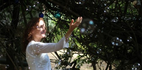 תמונה זו מציגה טיפול בחרדות של אישה עם שיער אדום עומדת מתחת לעצים ומושיטה את ידה לכיוון בועות סבון.